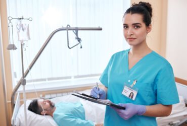 Portrait of serious young nurse in surgical gloves making notes about patients health while filling medical card in hospital